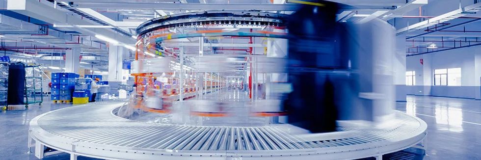 Fotografie din articolul PR electronics: instalație industrială cu bandă transportoare circulară cu role și recipiente în mișcare.